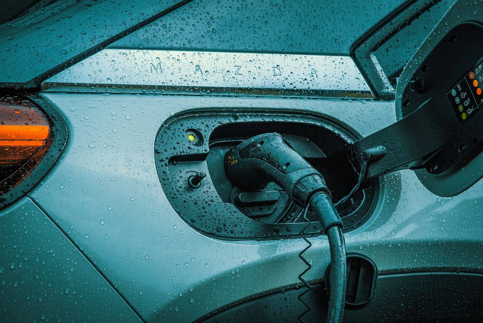 Close-up of a rain-covered car with a charging connector plugged into its port, lit in teal and orange